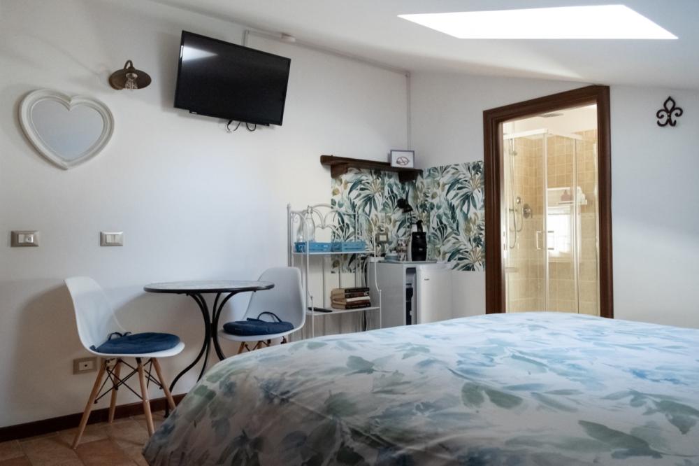 Attic double bedroom Orchidea at B&B C'era una volta with blue leaf-patterned bedding, a small table with two white chairs, an in-room coffee station, and an open door to the private bathroom with a glass shower.