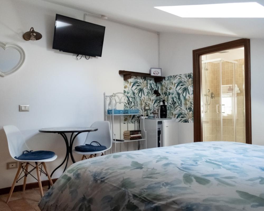 Attic double bedroom Orchidea at B&amp;B C'era una volta with blue leaf-patterned bedding, a small table with two white chairs, an in-room coffee station, and an open door to the private bathroom with a glass shower.