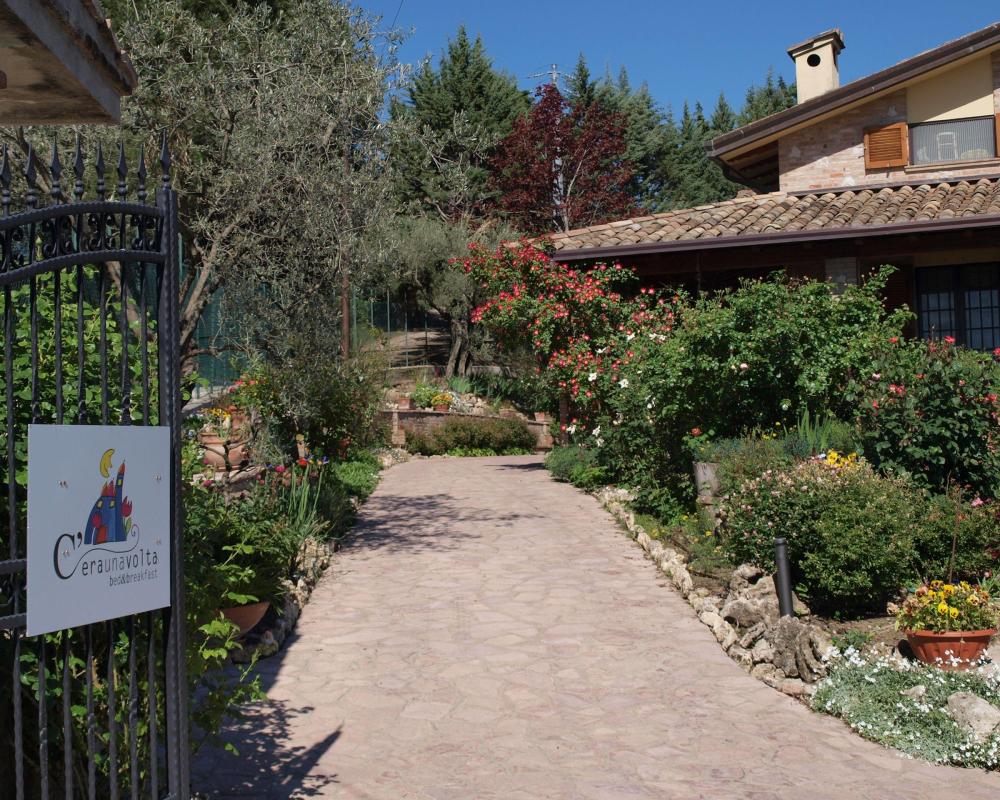 La facciata in pietra del B&amp;B C'era una volta vista dal vialetto pavimentato del giardino, circondata da alberi verdi, piante e un limpido cielo azzurro.