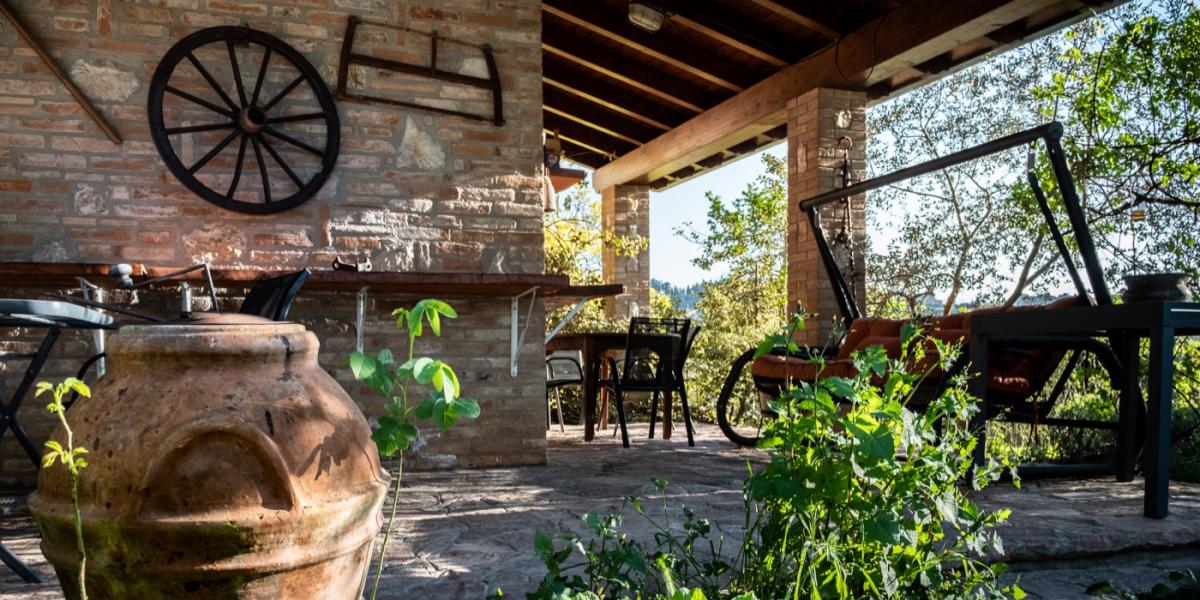 Accogliente portico esterno e angolo relax in giardino del B&amp;B C'era una volta, con tavolino e sedie accanto a una parete in mattoni decorata con una vecchia ruota di carro e attrezzi rustici.