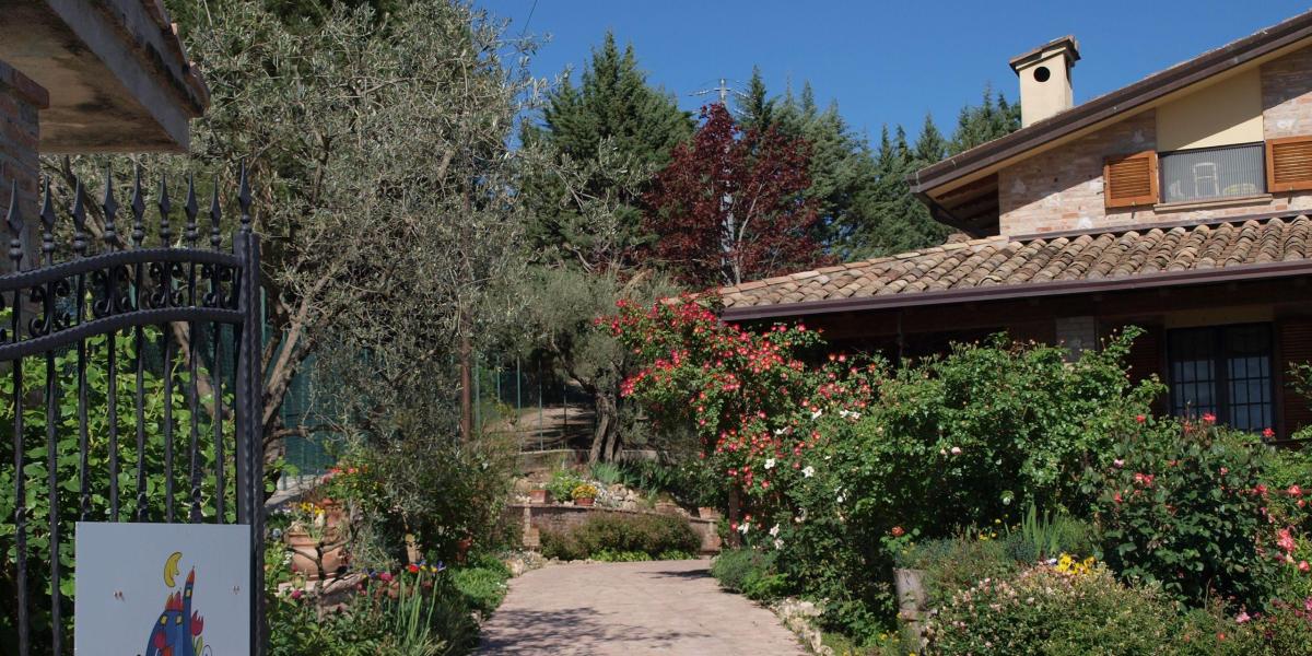 La facciata in pietra del B&amp;B C'era una volta vista dal vialetto pavimentato del giardino, circondata da alberi verdi, piante e un limpido cielo azzurro.